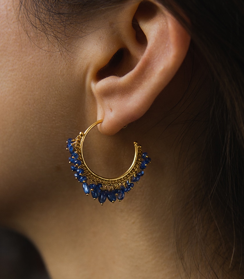 A close up if a model wearing a pair of blue sapphire beaded hoop earrings in gold vermeil