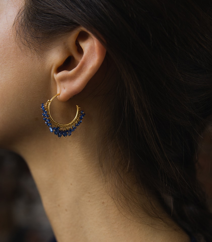 A dark haired model wearing a pair of blue sapphire hoop earrings in gold plated silver