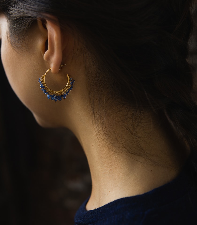 A close up of a dark haired model wearing a pair of blue sapphire beaded hoop earrings in gold vermeil