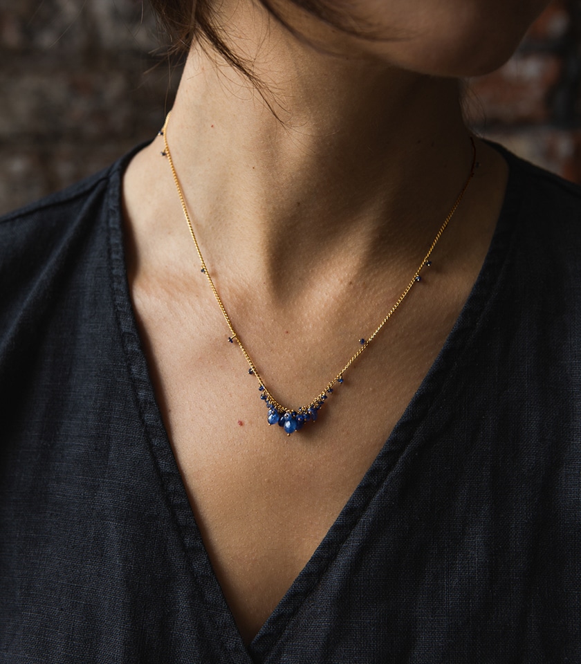 A model's neck and shoulders wearing a blue sapphire bead necklace on gold vermeil chain and a black linen wrap top