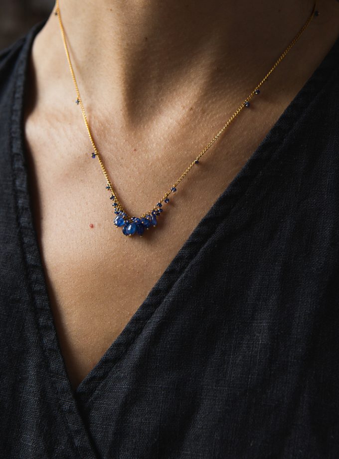 A model's neck and shoulders wearing a blue sapphire bead necklace on gold vermeil chain and a black linen top