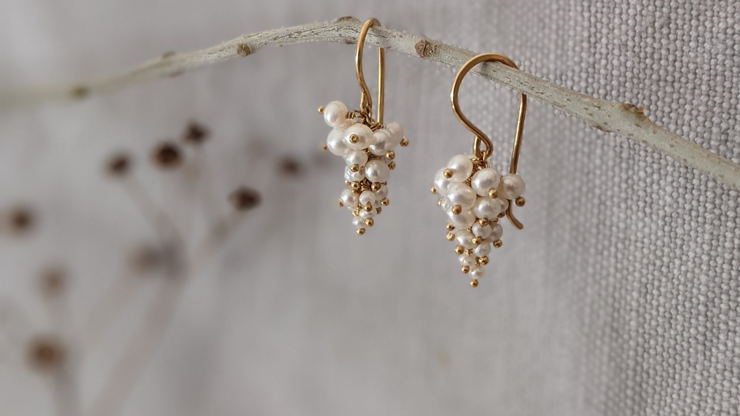 A pair of grape shaped pearl and gold vermeil earrings on a twig in front of a grey linen backdrop